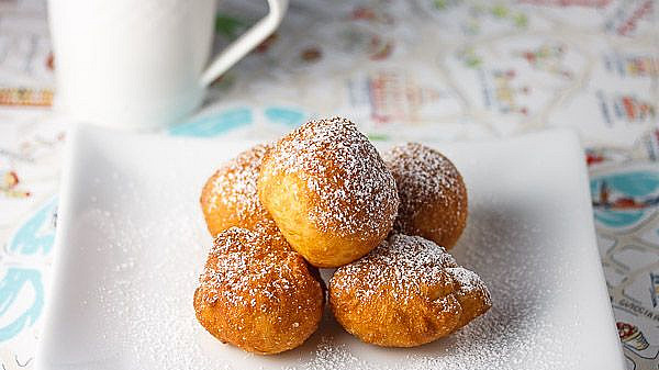 A white plate holding six golden-brown fritters dusted with powdered sugar, accompanied by a white coffee cup in the background, set on a patterned tablecloth.