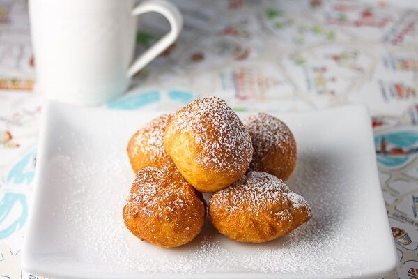 A white plate holding six golden-brown fritters dusted with powdered sugar, accompanied by a white coffee cup in the background, set on a patterned tablecloth.
