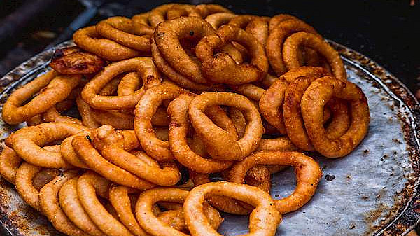 A close-up image shows a pile of freshly cooked, golden-brown ring-shaped fritters on a metal plate. The fritters appear crispy and are visibly coated in seasoned batter. The background is slightly blurred, focusing attention on the food.