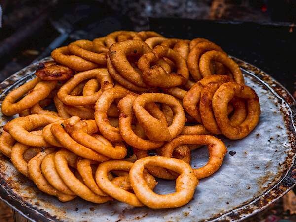 A close-up image shows a pile of freshly cooked, golden-brown ring-shaped fritters on a metal plate. The fritters appear crispy and are visibly coated in seasoned batter. The background is slightly blurred, focusing attention on the food.