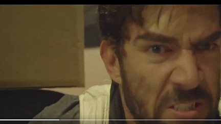 A man with a beard and intense facial expression appears angry or frustrated. He is indoors, and part of a brown object is visible to his left. The image is slightly blurred.
