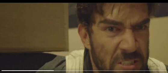 A man with a beard and intense facial expression appears angry or frustrated. He is indoors, and part of a brown object is visible to his left. The image is slightly blurred.