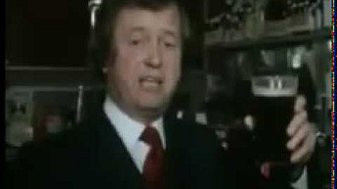 A man in a dark suit and red tie is holding up a pint of dark beer in a bar or pub setting. He appears to be in mid-speech, with shelves and bottles visible in the background.