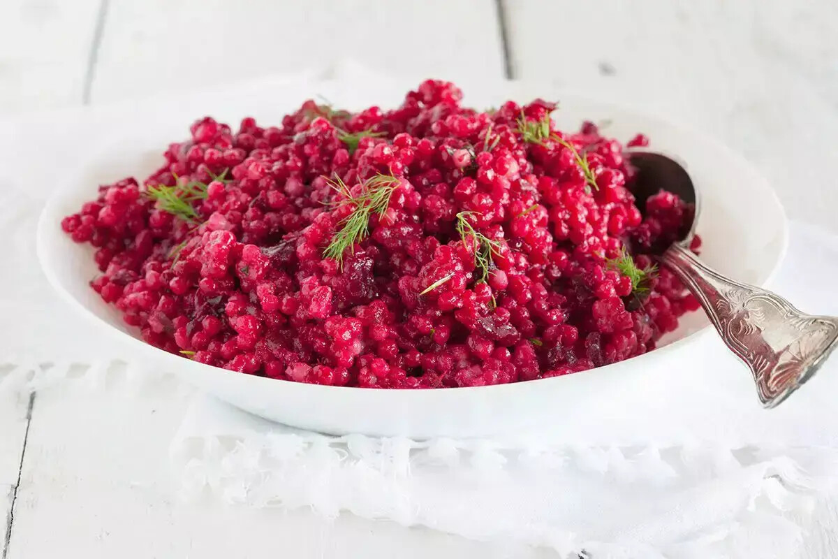 A white oval dish filled with bright red beetroot salad garnished with dill, placed on a white wooden surface. a silver spoon rests inside the dish to the right.