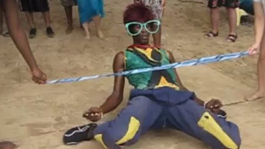 A person wearing large green sunglasses and a colorful outfit is performing a limbo dance under a blue and white stick on a sandy surface. Several people are standing around, watching the performance.