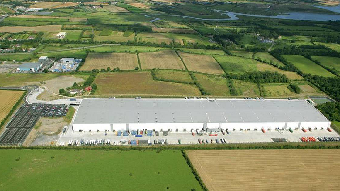 Aerial view of a large industrial warehouse set in a rural area surrounded by fields and farmland. The warehouse has a white roof, and numerous trailers and containers are parked around it. A river and various other buildings are visible in the distance.