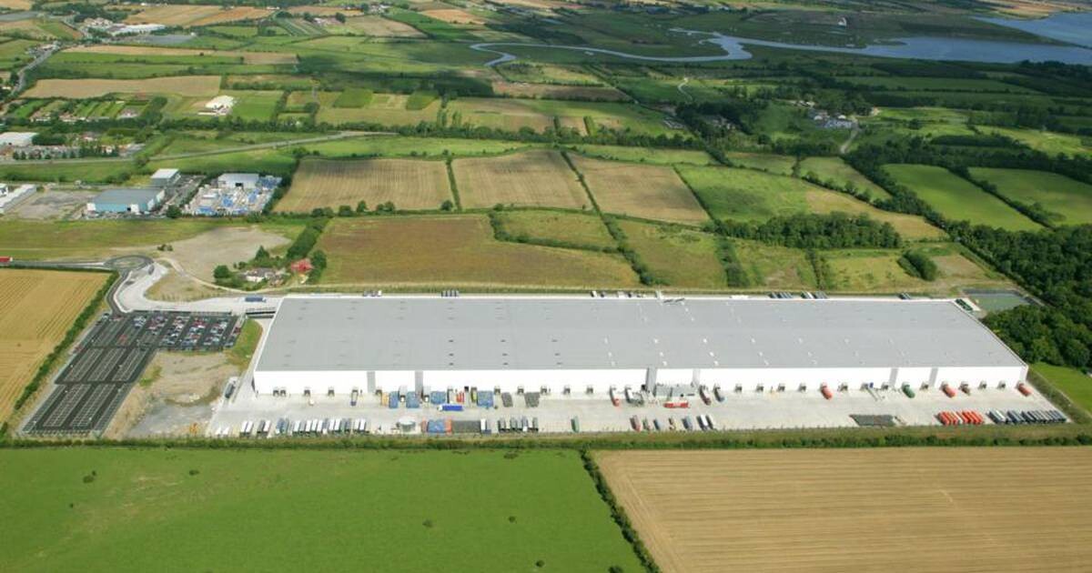 Aerial view of a large industrial warehouse set in a rural area surrounded by fields and farmland. The warehouse has a white roof, and numerous trailers and containers are parked around it. A river and various other buildings are visible in the distance.