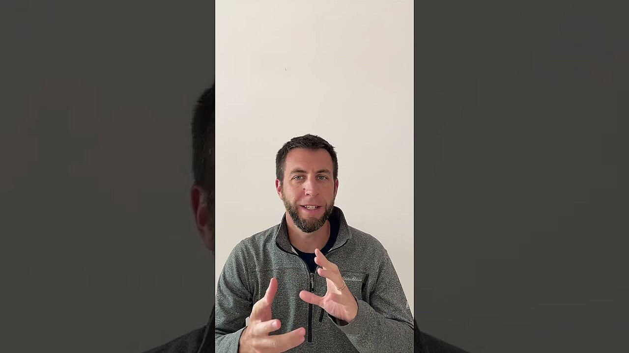 A person with short hair and a beard is seated against a plain white background. They are wearing a gray zip-up jacket and gesturing with their hands as if explaining something. The image has a portrait orientation.