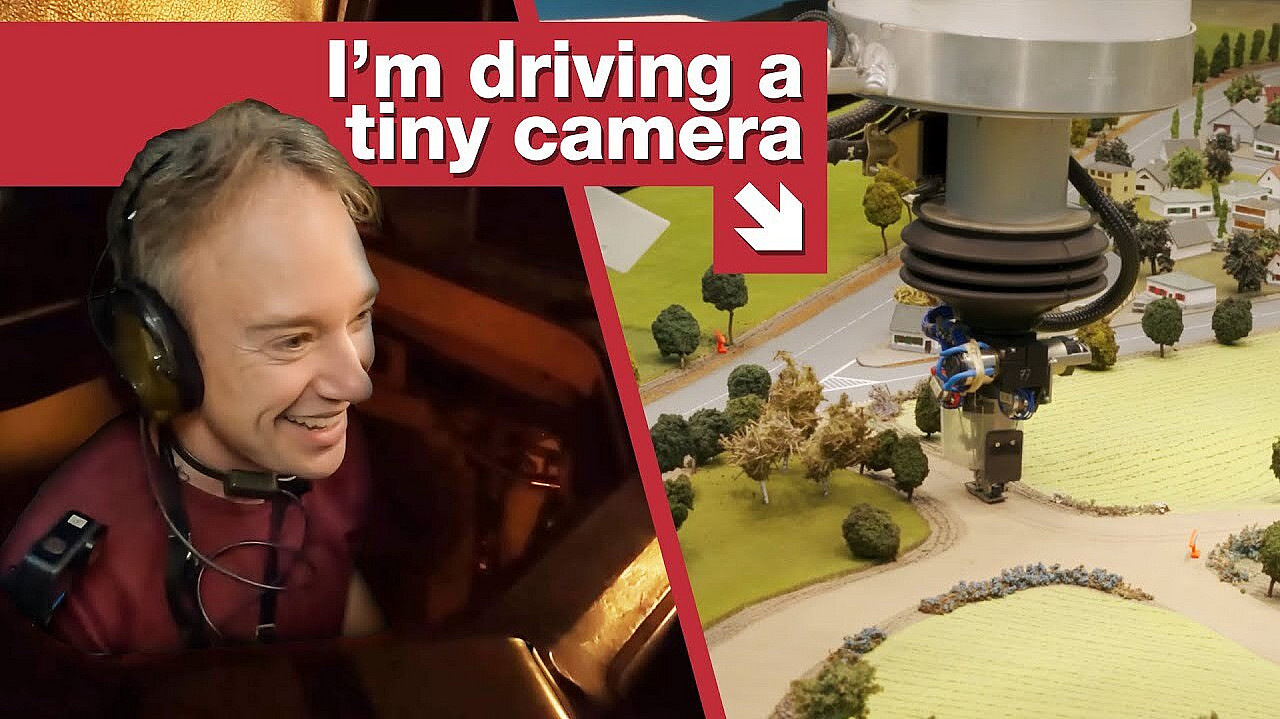 A person is smiling and wearing a headset on the left side of the image. On the right side, a small camera attached to a mechanical arm is recording a miniature model landscape with trees and fields. The text reads, "I'm driving a tiny camera.