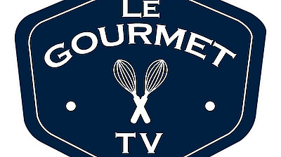 A dark blue hexagonal logo with a white outline. Inside, the text "Le GOURMET TV" is written in white, with two crossed whisks between "Le" and "GOURMET," and two small white dots flanking these elements.