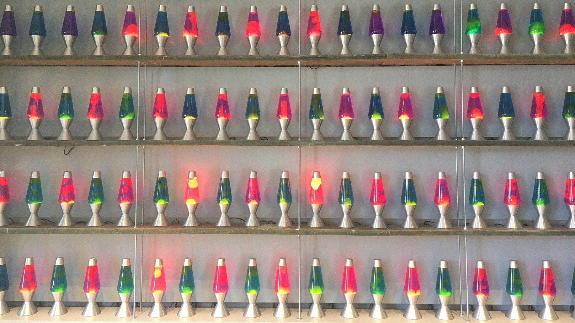A display of multiple rows of lava lamps, each in different colors, arranged on shelves. The lava lamps have a variety of wax colors within, creating a vibrant and colorful scene against a plain wall background.