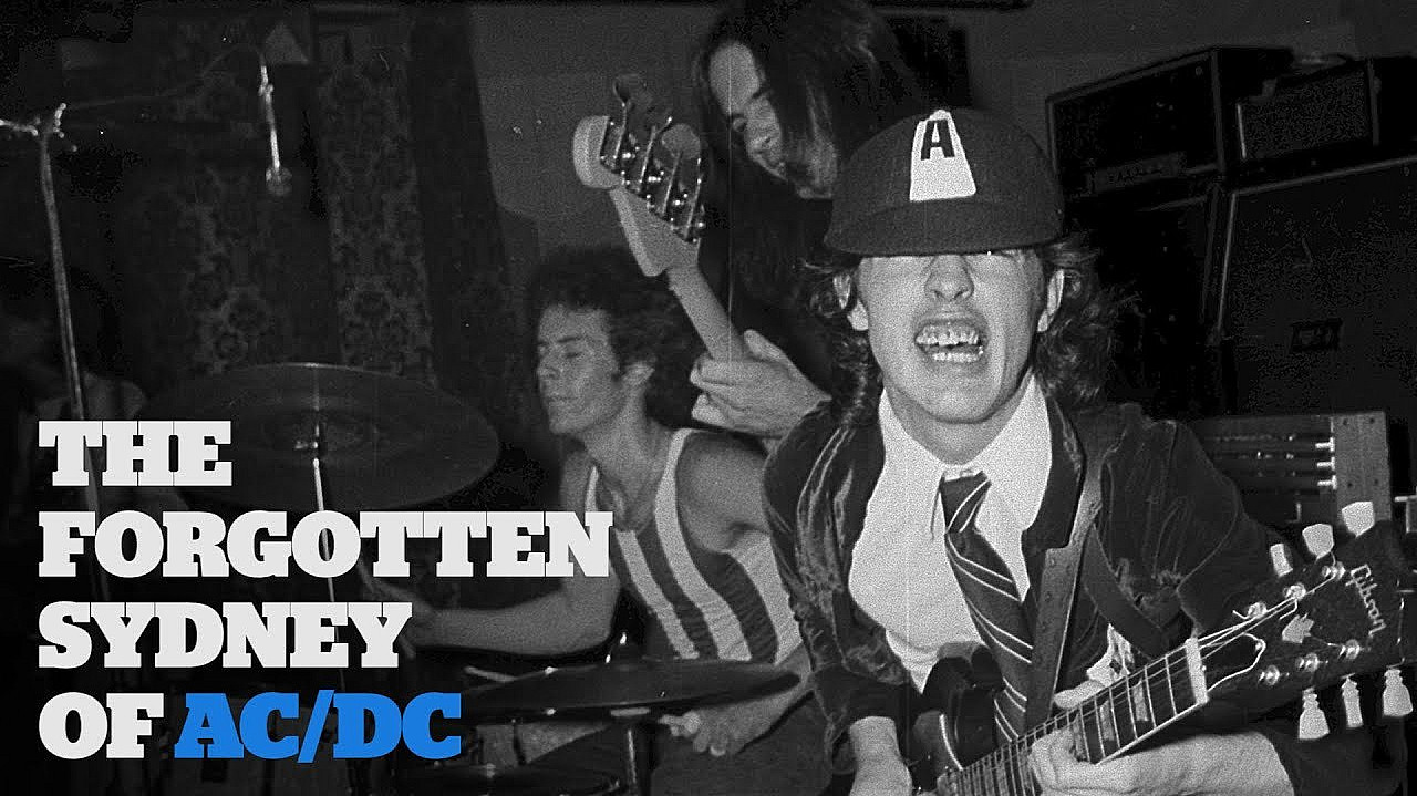 Black and white photo shows a band performing. The foreground features a musician wearing a cap with the letter "A" while playing guitar. Another musician is playing a guitar in the background, and a third is on drums. Text on the left reads "The Forgotten Sydney of AC/DC.