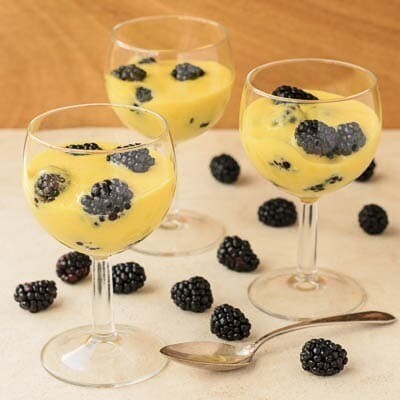 Three glasses of custard topped with blackberries on a light wood surface, accompanied by extra blackberries and a spoon.