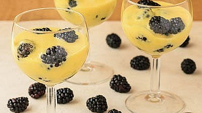 Three glasses of custard topped with blackberries on a light wood surface, accompanied by extra blackberries and a spoon.