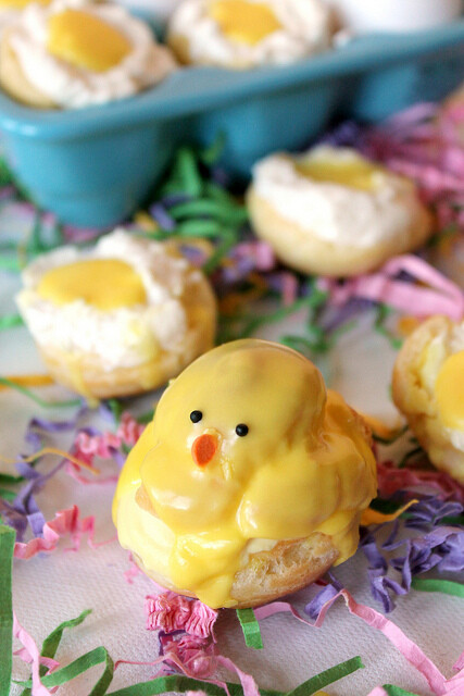 A decorative easter treat shaped like a chick, made from a doughnut topped with yellow icing and candy features, displayed on colourful shredded paper with other pastries in the background.