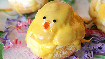 A decorative easter treat shaped like a chick, made from a doughnut topped with yellow icing and candy features, displayed on colourful shredded paper with other pastries in the background.