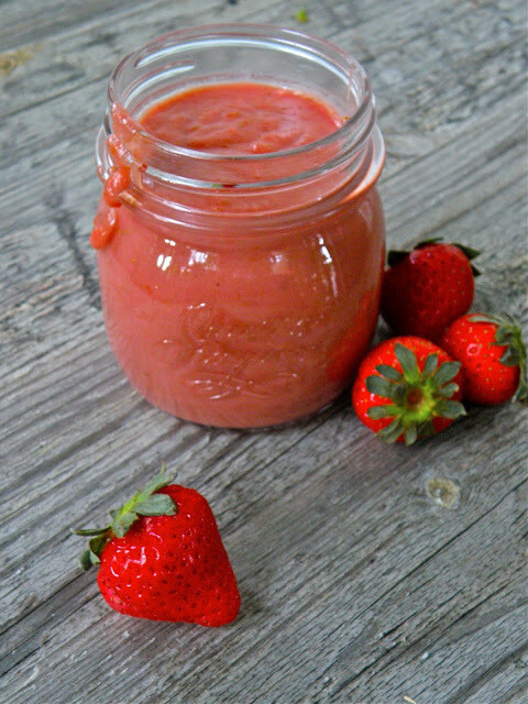A glass jar filled with strawberry curd on a wooden surface, surrounded by fresh strawberries.
