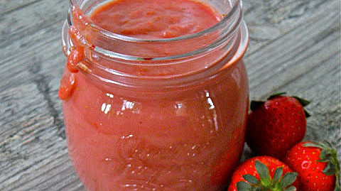 A glass jar filled with strawberry curd on a wooden surface, surrounded by fresh strawberries.