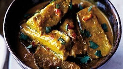 A bowl of aubergine curry, featuring large chunks of augbergine in a rich, golden sauce garnished with fresh cilantro, presented in a dark bowl against a light background.