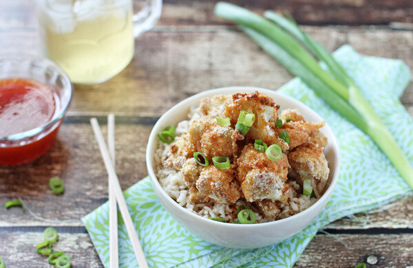 A bowl of crispy fried cauliflower over rice, garnished with green onions, next to chopsticks and dipping sauce on a rustic wooden table.