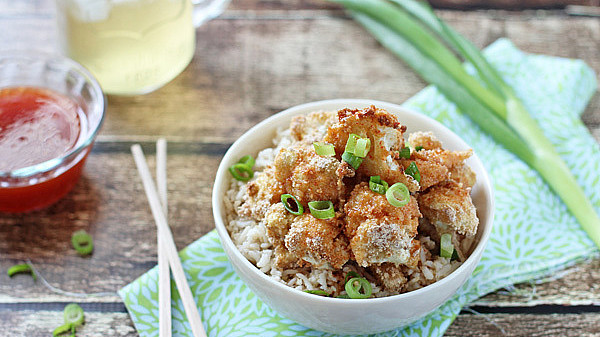 A bowl of crispy fried cauliflower over rice, garnished with green onions, next to chopsticks and dipping sauce on a rustic wooden table.
