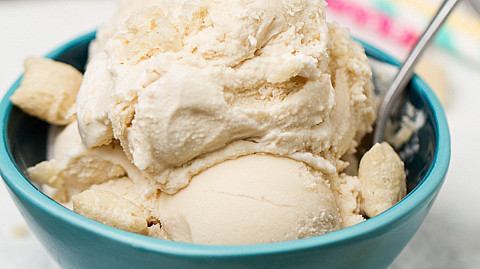 Two scoops of vanilla ice cream with cookie pieces served in a turquoise bowl with a spoon, placed on a wooden surface with colorful straws in the background.