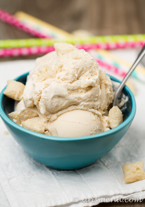 Two scoops of vanilla ice cream with cookie pieces served in a turquoise bowl with a spoon, placed on a wooden surface with colorful straws in the background.