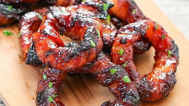 Grilled bacon-wrapped onion rings on a wooden cutting board, garnished with chopped parsley, displaying a shiny, caramelized exterior.