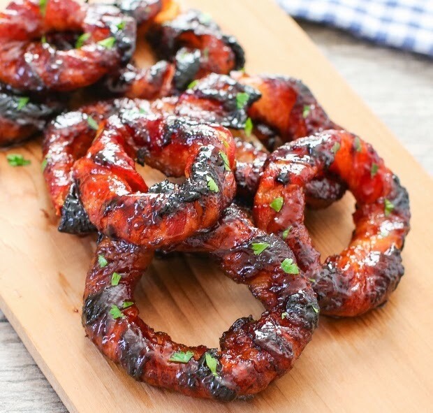 Grilled bacon-wrapped onion rings on a wooden cutting board, garnished with chopped parsley, displaying a shiny, caramelized exterior.