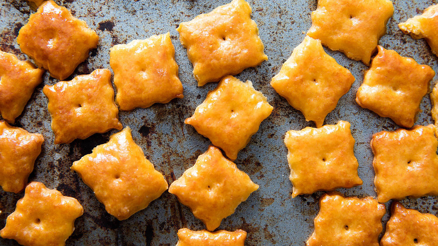 Baked square cheese crackers with a golden-brown hue, scattered on a rustic, spotted baking tray.