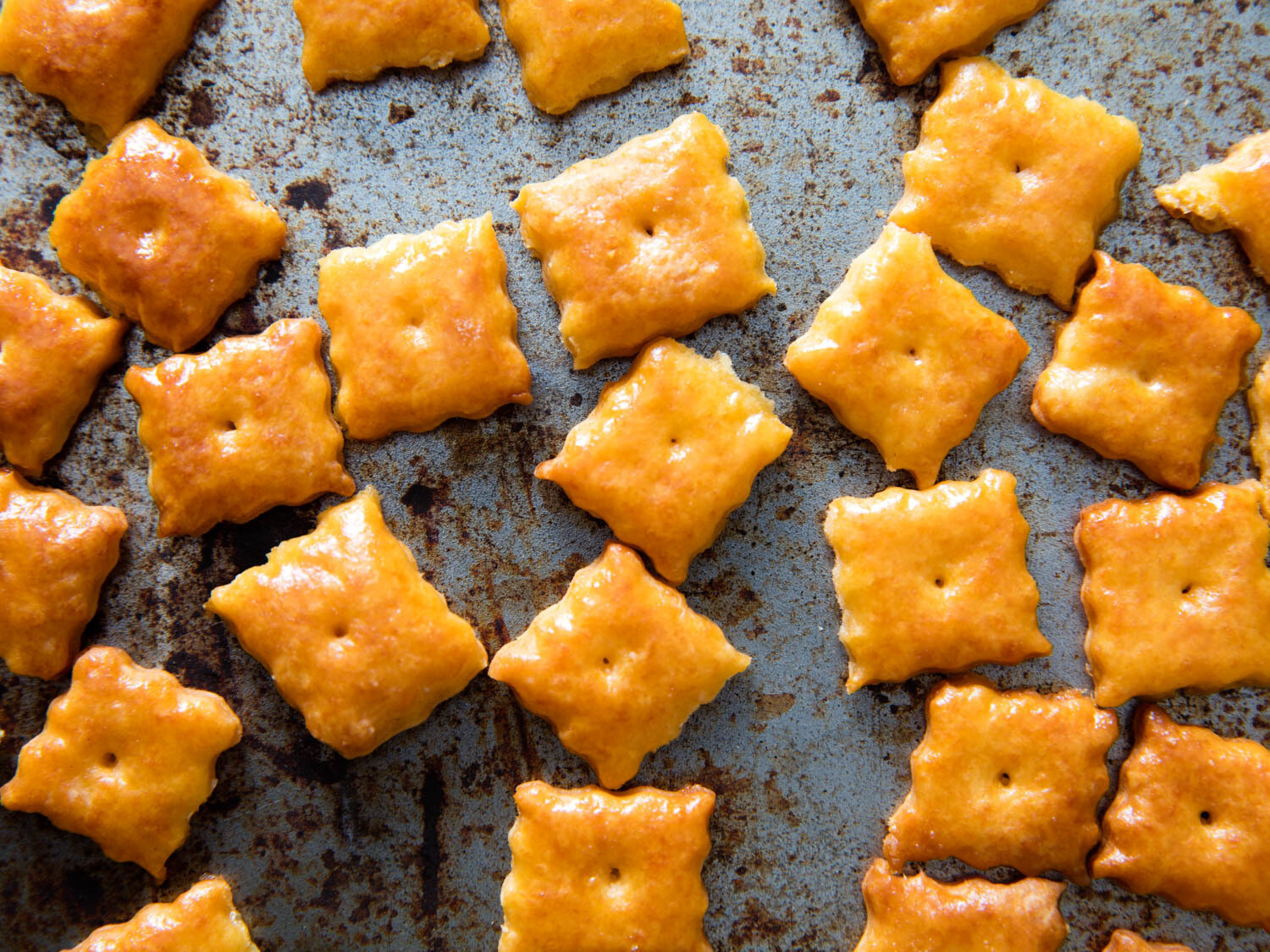 Baked square cheese crackers with a golden-brown hue, scattered on a rustic, spotted baking tray.