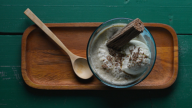 A glass bowl with vanilla affogato, topped with a flake chocolate bar and grated chocolate, sits on a wooden tray with a wooden spoon against a green wooden background.