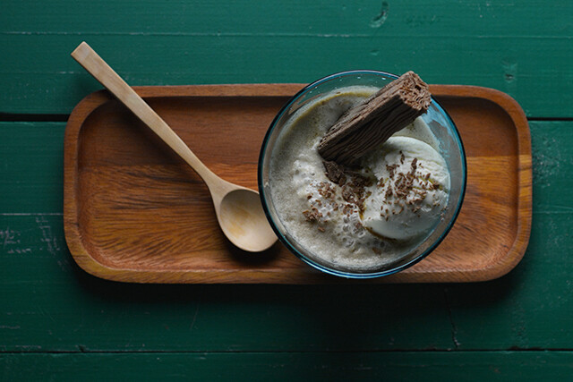A glass bowl with vanilla affogato, topped with a flake chocolate bar and grated chocolate, sits on a wooden tray with a wooden spoon against a green wooden background.