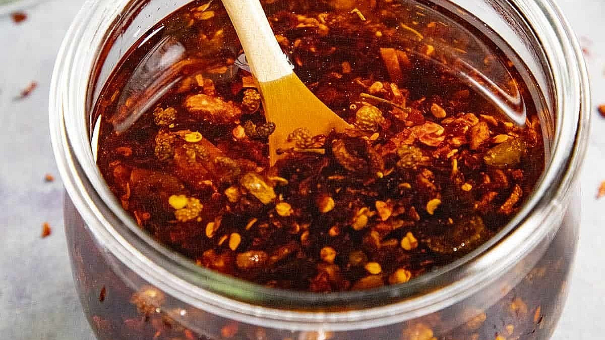 A glass jar filled with chili oil, featuring flakes of chili pepper. a wooden spoon is inserted into the oil. the jar is on a light surface with a green chili pepper and glass jar in the background.
