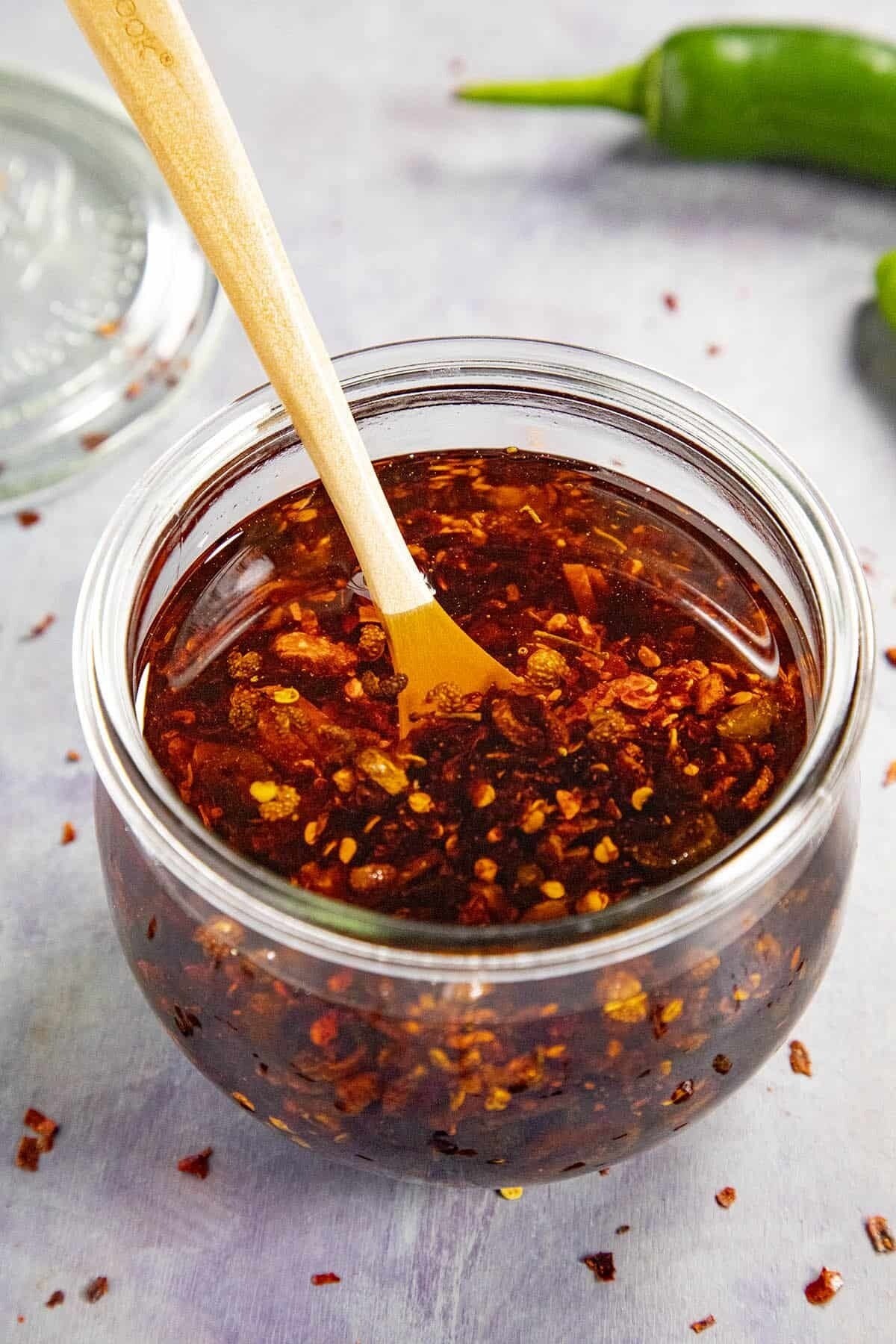 A glass jar filled with chili oil, featuring flakes of chili pepper. a wooden spoon is inserted into the oil. the jar is on a light surface with a green chili pepper and glass jar in the background.
