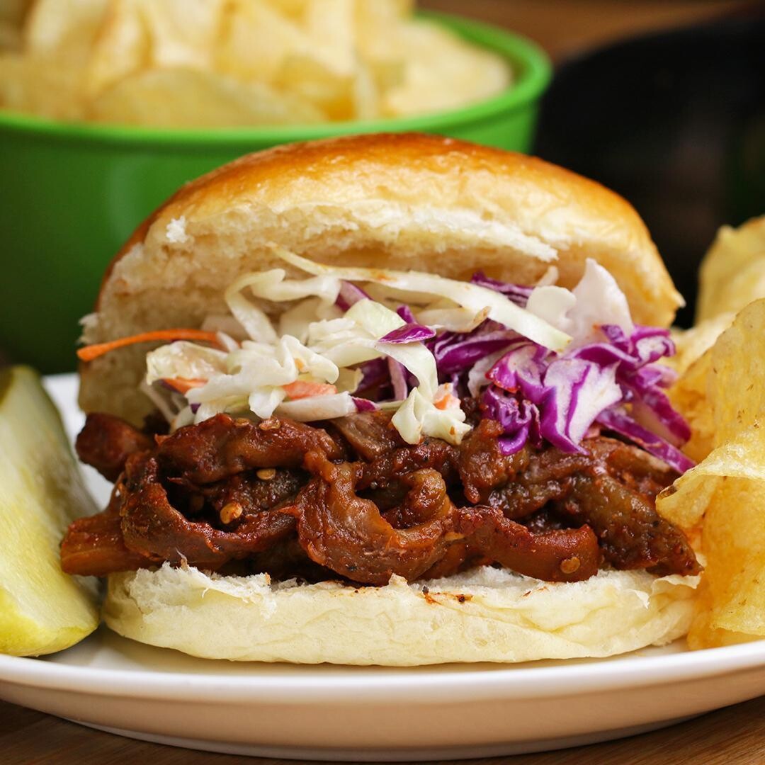 A bbq pulled eggplant sandwich with coleslaw and red cabbage on a bun, accompanied by a side of potato chips and a small green bowl in the background.