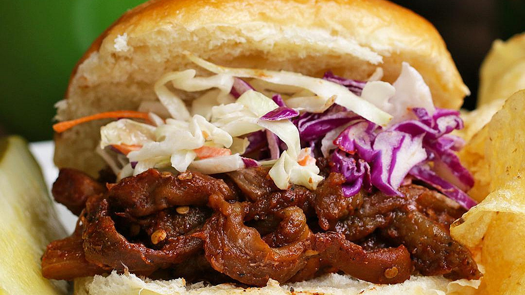 A bbq pulled eggplant sandwich with coleslaw and red cabbage on a bun, accompanied by a side of potato chips and a small green bowl in the background.