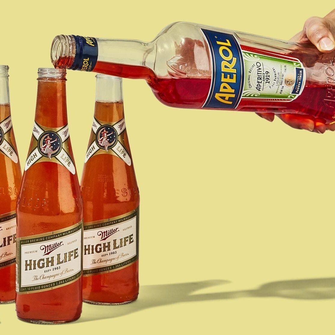 A hand pours Aperol into three Miller High life beer bottles against a yellow background, suggesting a cocktail preparation.