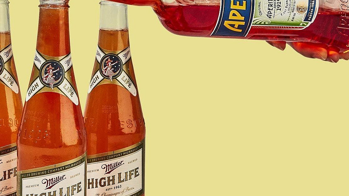 A hand pours Aperol into three Miller High life beer bottles against a yellow background, suggesting a cocktail preparation.