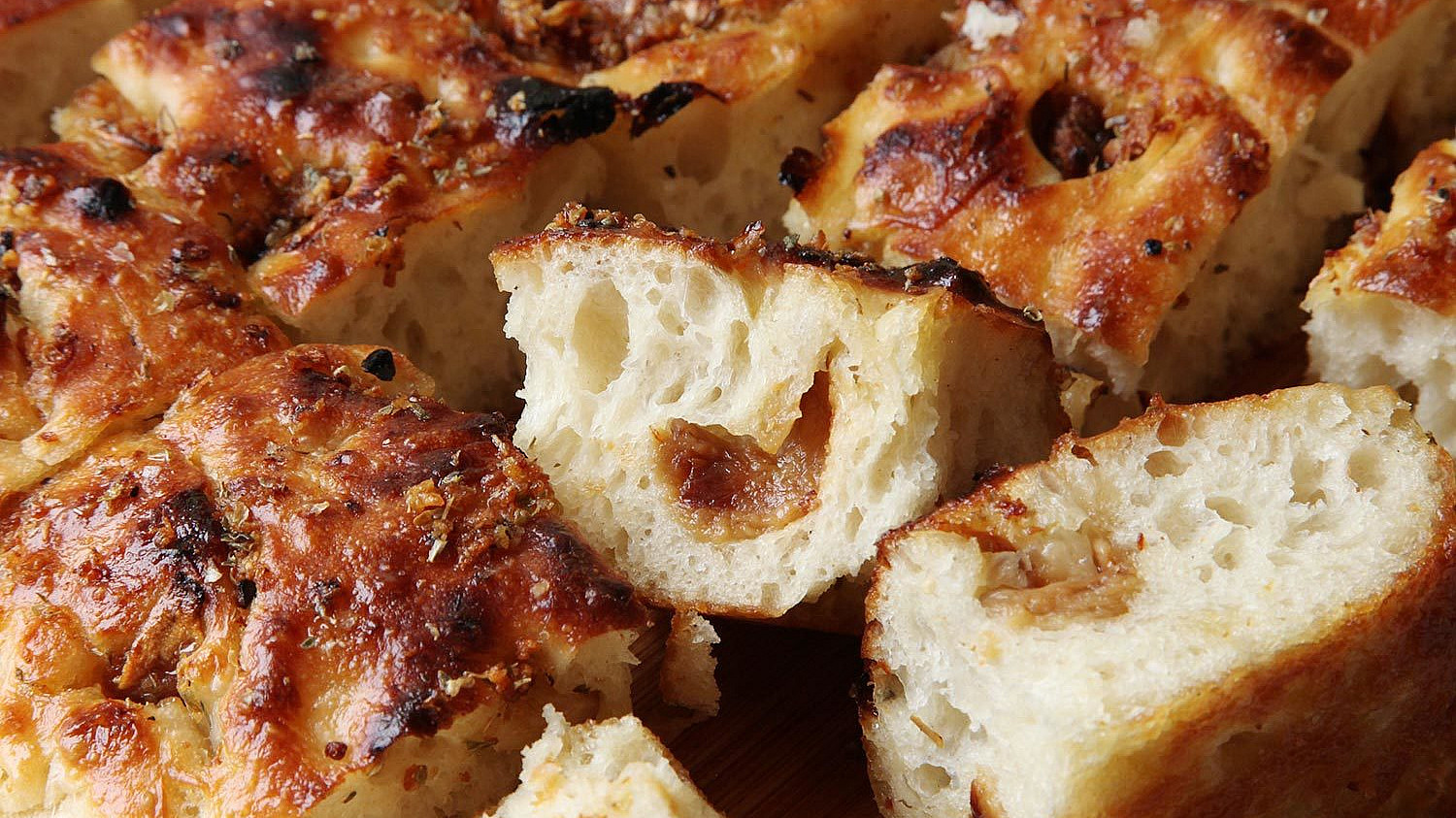 Close-up of a sliced focaccia bread, displaying its golden-brown, crispy crust and soft, airy interior. The bread is seasoned and appears to be infused with caramelized onions or a similar ingredient, evident from the visible bits and the coloration.
