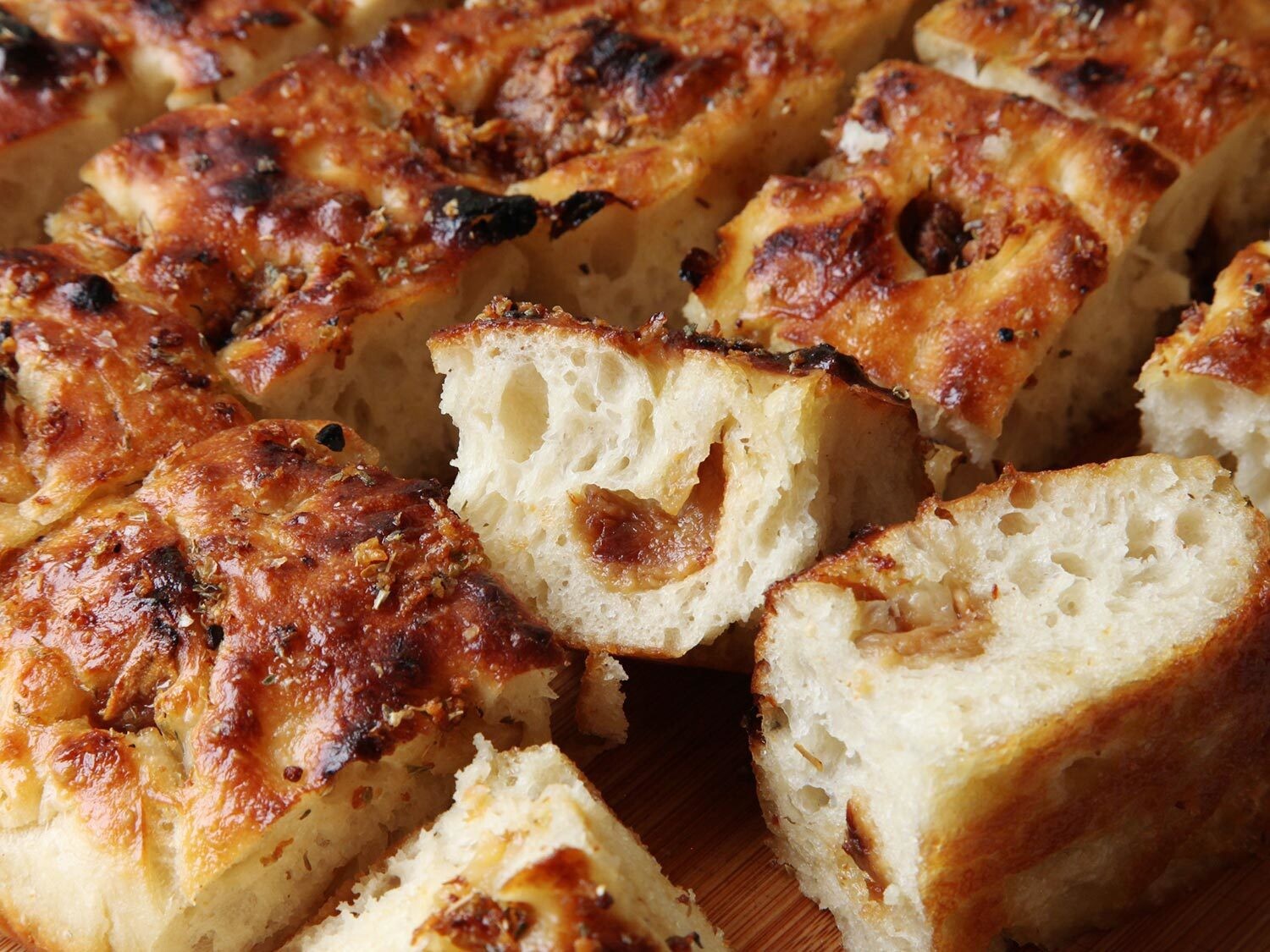 Close-up of a sliced focaccia bread, displaying its golden-brown, crispy crust and soft, airy interior. The bread is seasoned and appears to be infused with caramelized onions or a similar ingredient, evident from the visible bits and the coloration.