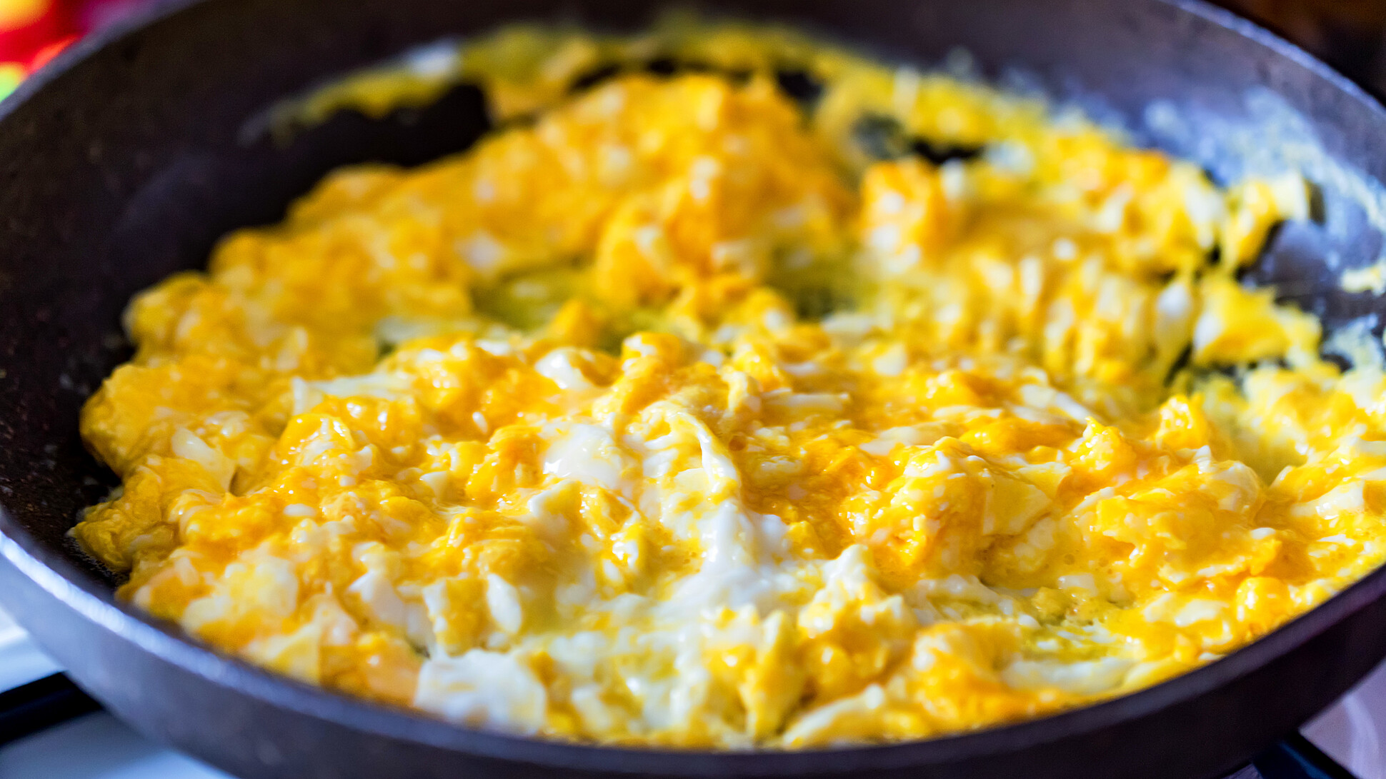 A pan on a stovetop contains scrambled eggs that are partially cooked. The eggs appear to be soft and slightly runny with a bright yellow color. There are small patches of white, indicating the presence of egg whites.