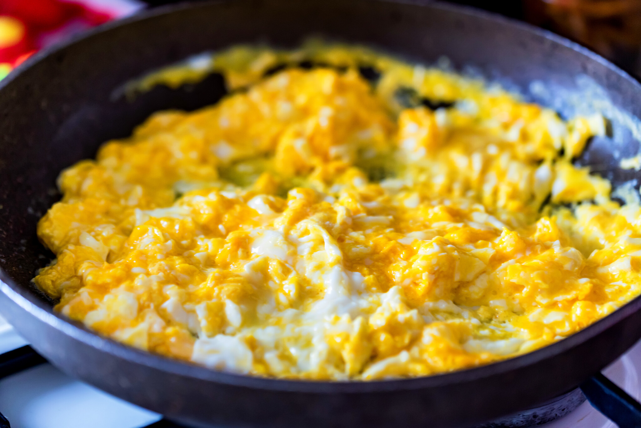 A pan on a stovetop contains scrambled eggs that are partially cooked. The eggs appear to be soft and slightly runny with a bright yellow color. There are small patches of white, indicating the presence of egg whites.