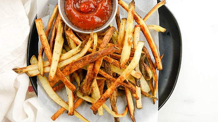 A plate of thinly sliced, crispy homemade fries is served with a small dish of ketchup. The fries are arranged on parchment paper with a black plate underneath. The background includes a white cloth and a glimpse of a red-striped towel.