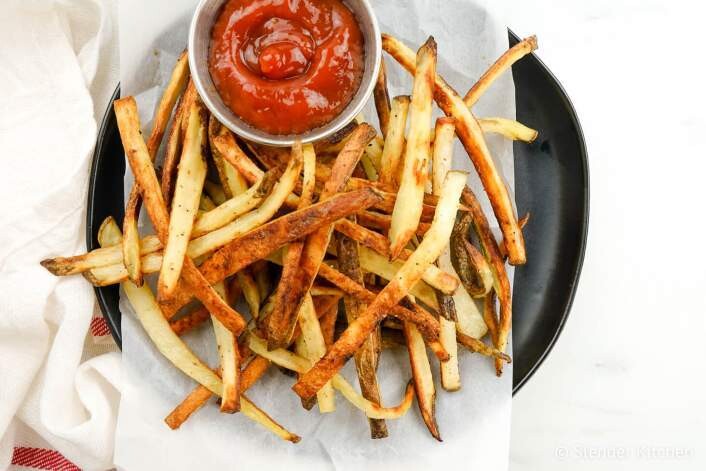 A plate of thinly sliced, crispy homemade fries is served with a small dish of ketchup. The fries are arranged on parchment paper with a black plate underneath. The background includes a white cloth and a glimpse of a red-striped towel.