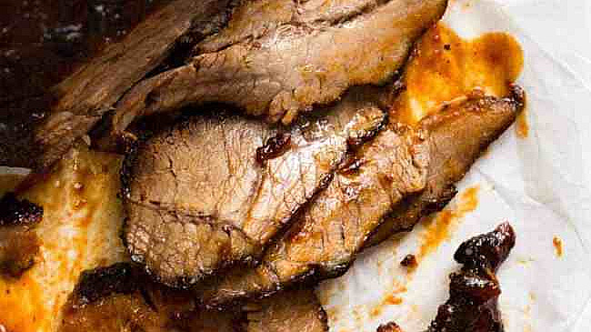 A close-up of sliced brisket on a piece of parchment paper. The meat is well-cooked, with a glazed, caramelized surface covered in barbecue sauce. Several slices are laid out, revealing a juicy and tender texture.