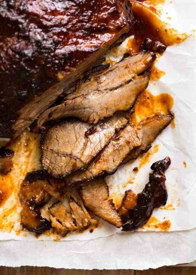 A close-up of sliced brisket on a piece of parchment paper. The meat is well-cooked, with a glazed, caramelized surface covered in barbecue sauce. Several slices are laid out, revealing a juicy and tender texture.
