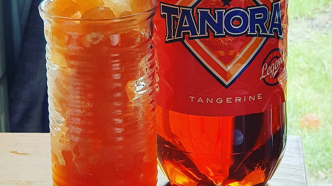 A glass with ice and orange-colored liquid stands next to a bottle of Tanora sparkling tangerine-flavored soda. A pink straw is inserted into the glass. The scene is set on a wooden surface with a hint of greenery in the background.