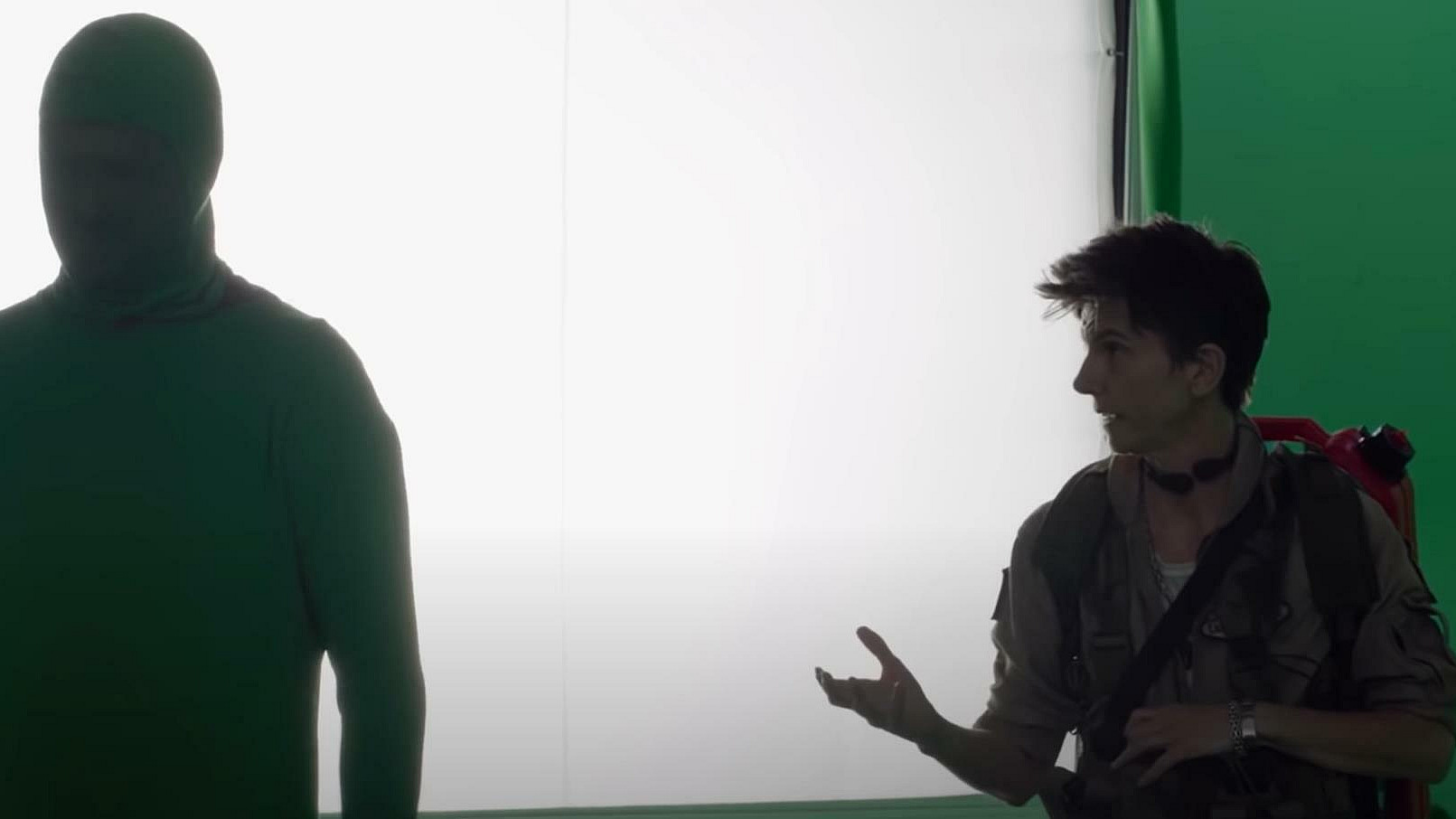 A person in a green full-body suit stands next to another person wearing a brown jacket and a backpack. The background includes green and white panels. They appear to be on a film or photography set.