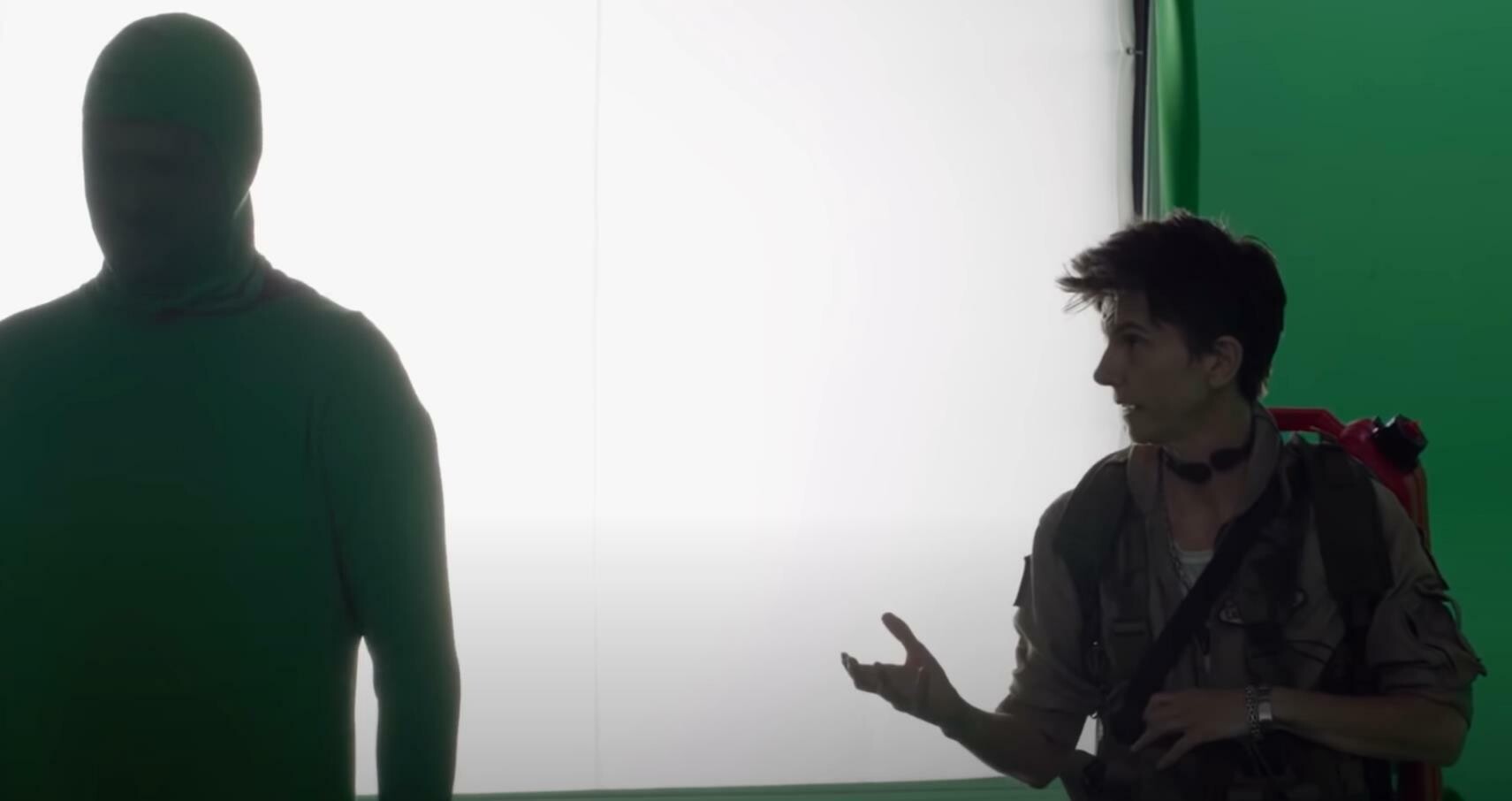 A person in a green full-body suit stands next to another person wearing a brown jacket and a backpack. The background includes green and white panels. They appear to be on a film or photography set.